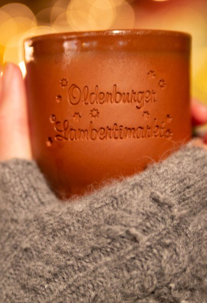 Eine Becher Glühwein mit der Aufschrift Oldenburger Lamberti-Markt wird in der Hand gehalten.