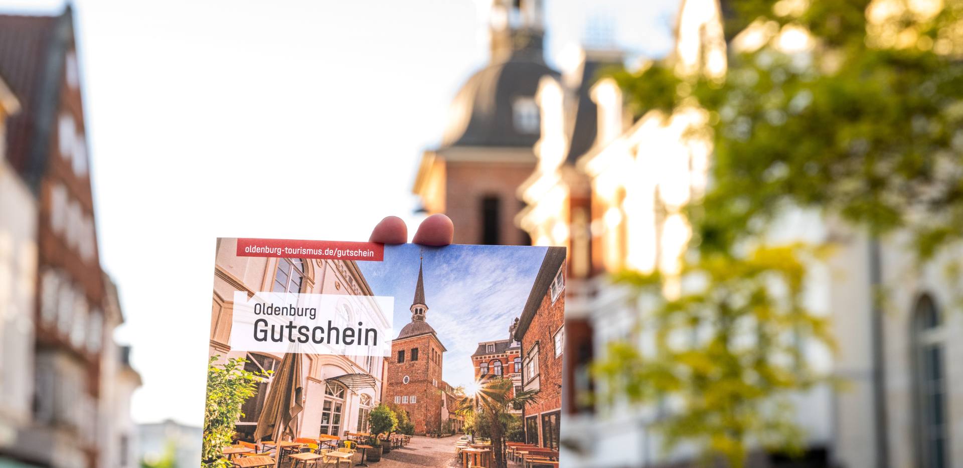 Eine Hand hält die Klappkarte des Oldenburg-Gutscheins in der Fußgängerzone mit dem Lappan im Hintergrund.