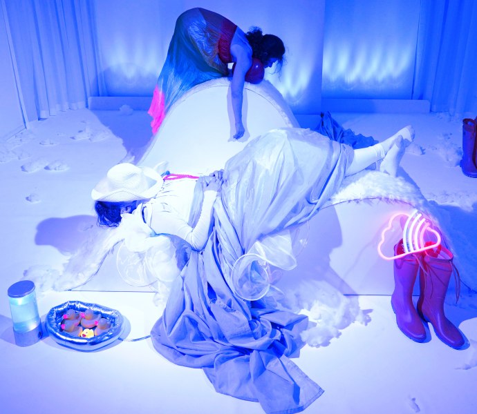 Zwei Schauspielerinnen in blaues Licht geh&uuml;llt, die eine schl&auml;ft scheinbar, die andere tr&auml;gt etwas.