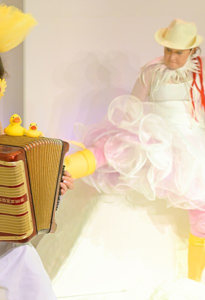 Zwei Schauspielerinnen, die eine spielt im Vordergrund Akkordeon, die andere scheint nach ihr zu treten. Beide haben viele gelbe Gegenst&auml;nde bei oder an sich.