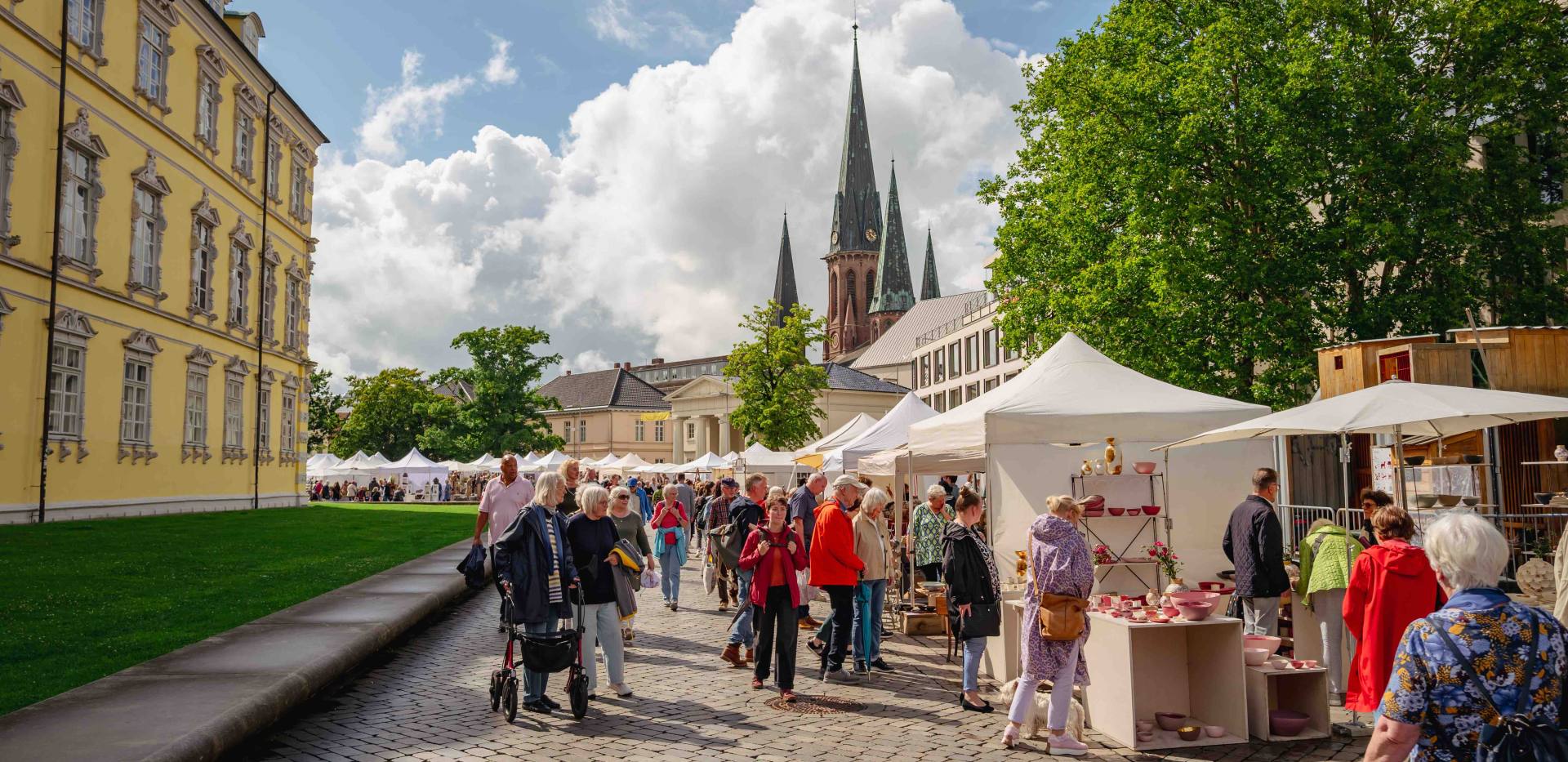 Links sieht man das gelbe Oldenburger Schloss und im fernen Hintergrund die Lambertikirche. Im Vordergrund sieht man verschiedene Menschen an den Keramikmarktst&auml;nden bummeln.