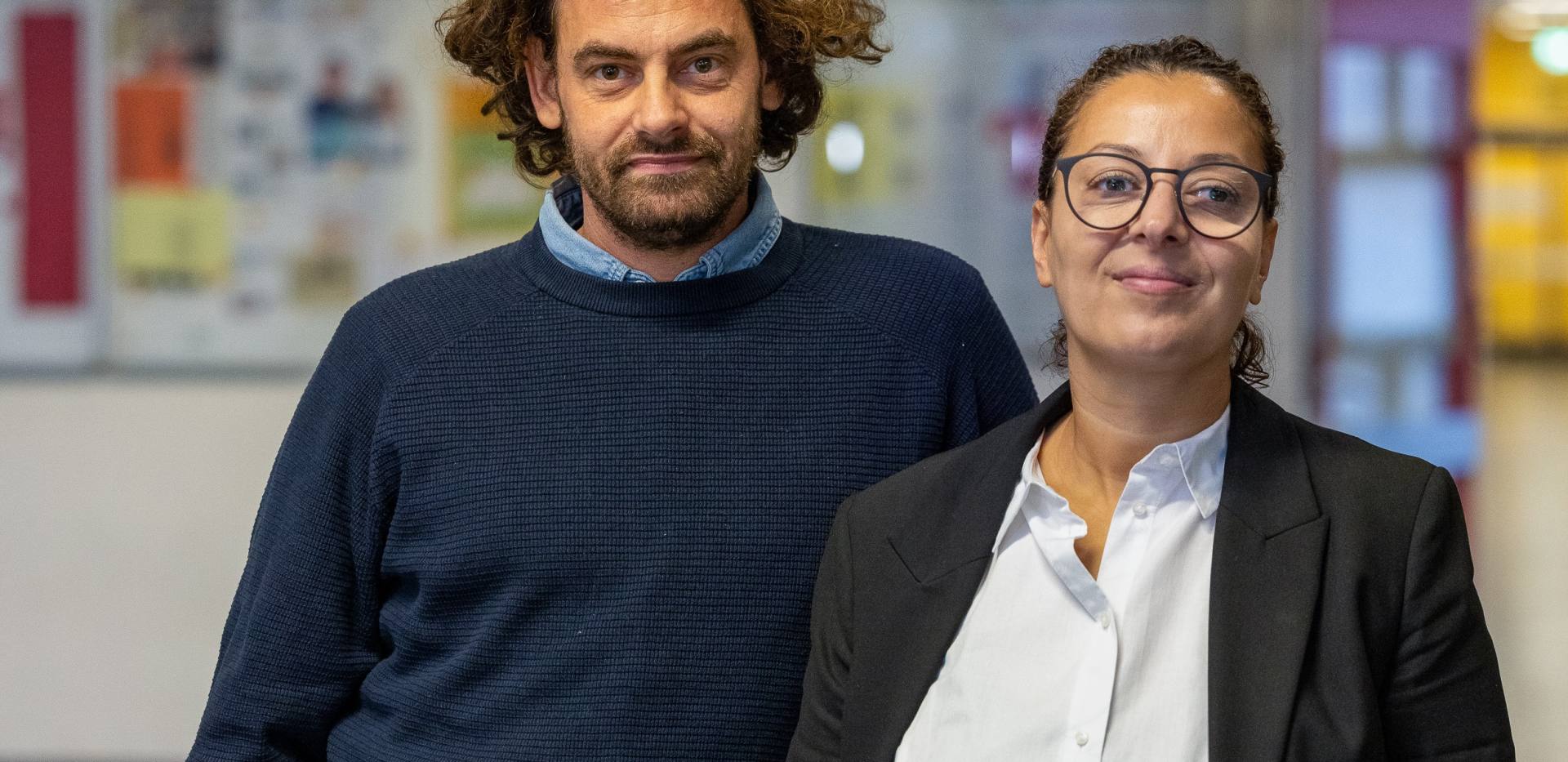 Brustbild von Shai Hoffmann und Jouanna Hassoun in frontaler Sicht. Beide stehen in einem Schulflur. Der Hintergrund ist unscharf, erkennbar sind - vermutlich von Schülerinnen angefertigte - Wandzeitungen an der Flurwand. Brustbild von Shai Hoffmann und Jouanna Hassoun in frontaler Sicht. Beide stehen in einem Schulflur. Der Hintergrund ist unscharf, erkennbar sind - vermutlich von Schülerinnen angefertigte - Wandzeitungen an der Flurwand.