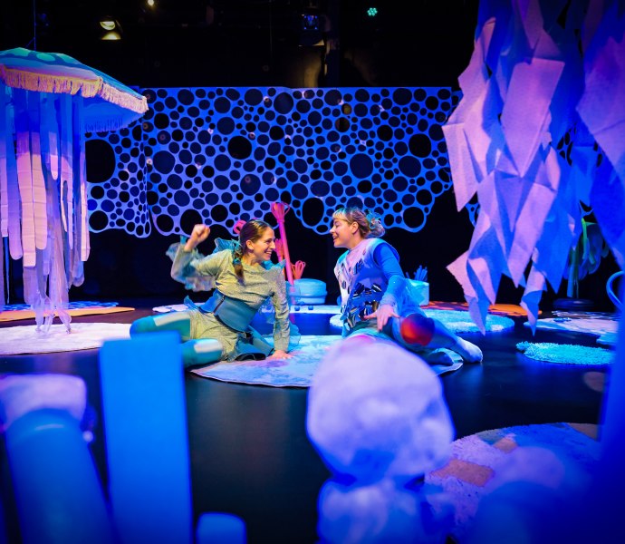 Zwei Performerinnen einander zugewandt auf einer B&uuml;hne in Unterwasseratmosph&auml;re