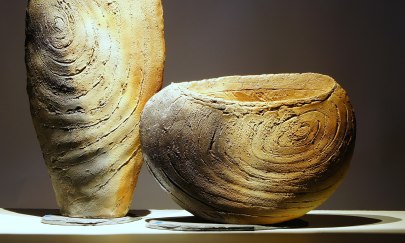Man sieht zwei große Keramische Gefäße mit rauer Oberfläche. Sie haben natürliche Erdtöne und stehen senkrecht auf einer glatten Oberfläche. Der Hintergrund ist sehr dunkel.