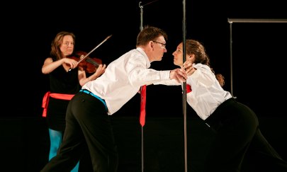 Zwei Spieler:innen stehen sich an einer Kleidungsstange gegenüber. Im Hintergrund spielt eine Person Geige. Zwei Spieler:innen stehen sich an einer Kleidungsstange gegenüber. Im Hintergrund spielt eine Person Geige.