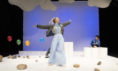 Schauspielerin tanzt auf B&uuml;hne, im Hintergrund schweben Ballons, die an Steinen festgemacht wurden, es ist eine andere Darstellerin zu sehen. Blauer Himmel und Wolken rahmen das B&uuml;hnenbild.