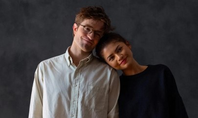 Auf dem Bild sind 2 Personen, die vor einem dunklen Hintergrund stehen. Die als Frau gelesene Person rechts lehnt sich an die linke Person, die als Mann gelesen wird. Beide Personen haben einen glücklichen / zufriedenen Gesichtsausdruck. Auf dem Bild sind 2 Personen, die vor einem dunklen Hintergrund stehen. Die als Frau gelesene Person rechts lehnt sich an die linke Person, die als Mann gelesen wird. Beide Personen haben einen glücklichen / zufriedenen Gesichtsausdruck.