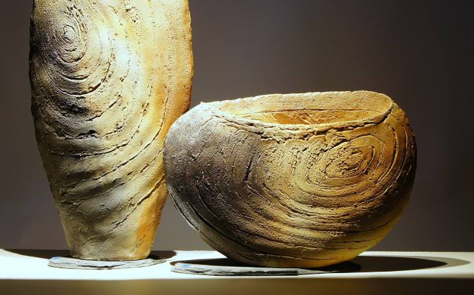 Man sieht zwei aus Ton hergestellte Gefäße die auf einer geraden Fläche stehen. Sie haben eine ungleichmäßige Oberfläche und Erdtöne. Der Hintergrund ist sehr dunkel.