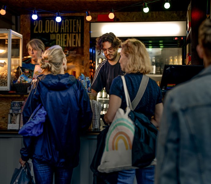 Perspektive auf den Kiosk am Platz, an dem mehrere Personen anstehen. Man Sieht zwei Personen, die im Kioskwagen Popcorn ausgeben, Getränke und Tickets.