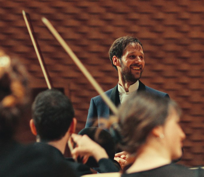 Ein lächelnder Mann steht vor einer Gruppe von Cello Spielern.