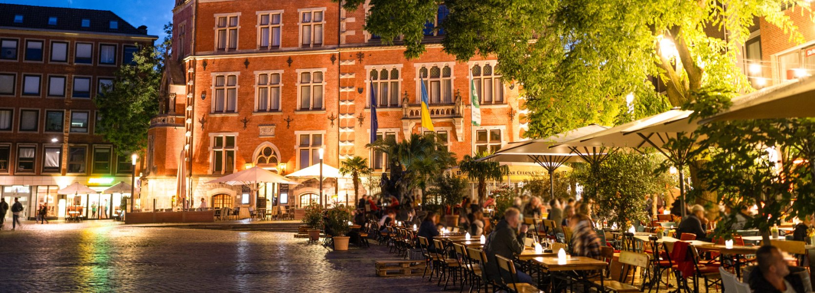 Am Oldenburger Rathausmarkt sitzen in der Abenddämmerung viele Menschen in den Lokalen vor dem beleuchteten Rathaus.