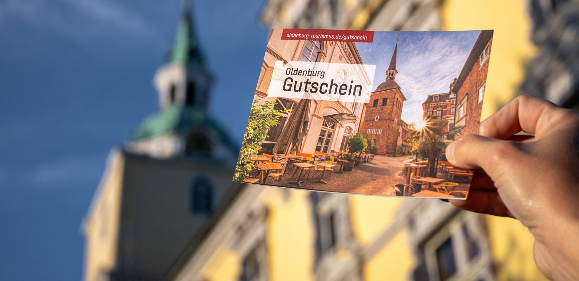 Eine Hand hält die Klappkarte des Oldenburg-Gutscheins vor dem Oldenburger Schloss