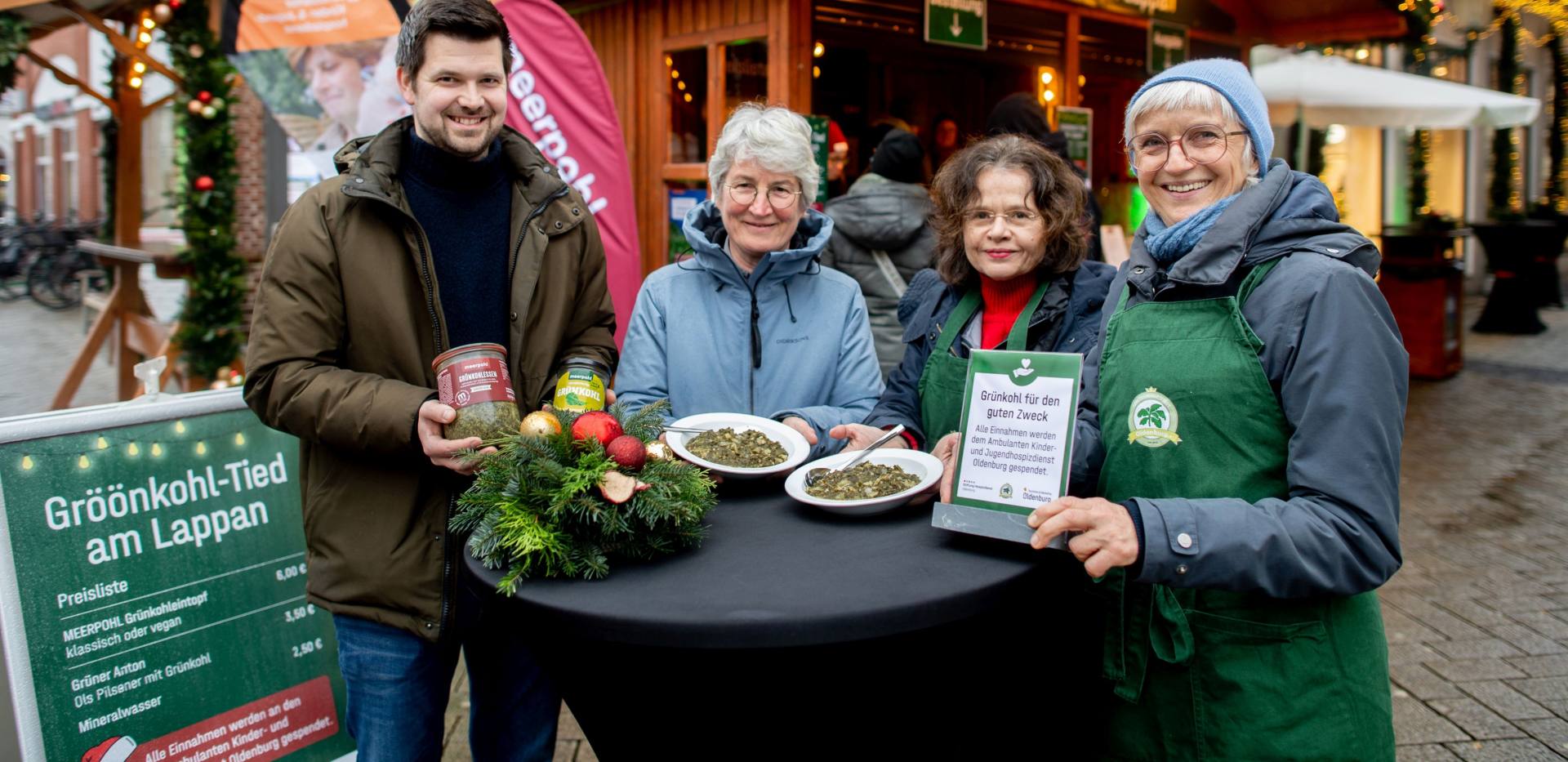 BU: Geschäftsführer Philipp Meerpohl, dessen Fleischerei den Grünkohleintopf für die „Gröönkohl-Tied“ vollständig spendete, gemeinsam mit Dr. Beate Jonas, Vorstandsvorsitzende des Hospizdienstes, und Beate Weidewitsch, Koordinatorin und Beraterin vor Ort. Am Stehtisch mit dem servierten Grünkohleintopf sind sie mit Silke Fennemann, Geschäftsführerin der OTM und Veranstalterin der Gröönkohl-Tied am Lappan, zu sehen. Die gesamten Erlöse gehen als Spende an den Hospizdienst.