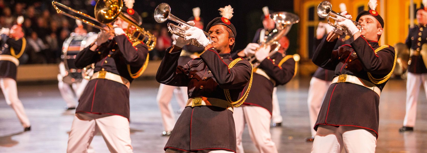 Ein internationales Blasorchester in prächtigen Uniformen begeistert mit schwungvoller Livemusik. Die Musiker performen im Gleichschritt mit Trompete und Posaune und vermitteln mit ihrer energiegeladenen Darbietung den einzigartigen Charakter der Musikparade. Die intensiven Farben und die Dynamik der Bewegung machen die Szene besonders eindrucksvoll.