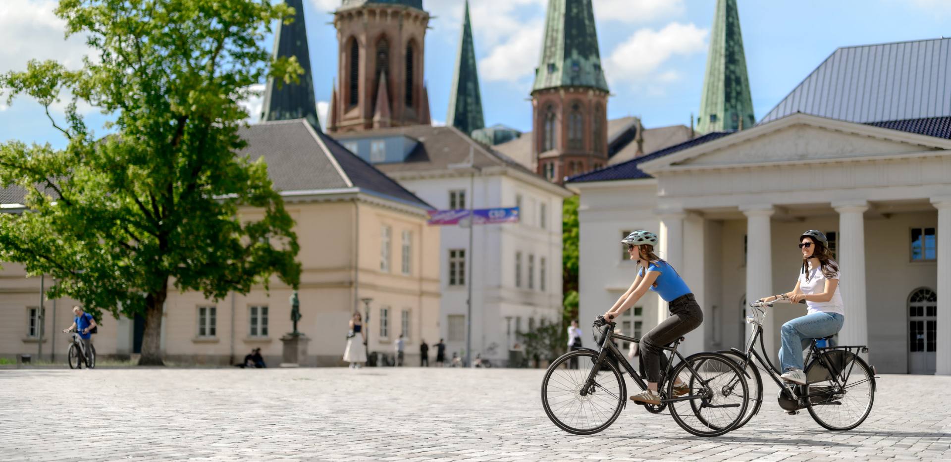 Radfahren Oldenburg Schlossplatz Zwei Radfahrerinnen auf dem Schlossplatz Oldenburg im Lamberti-Kirche und Schlosswache im Hintergrund.