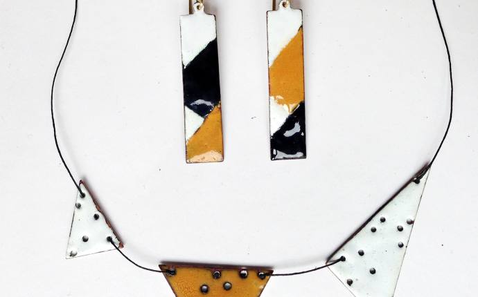 Man sieht rechteckige Ohrringe und eine Kette mit Dreiecken in den Farbe weiß, schwarz und orange die auf einem weißen Hintergrund liegen.