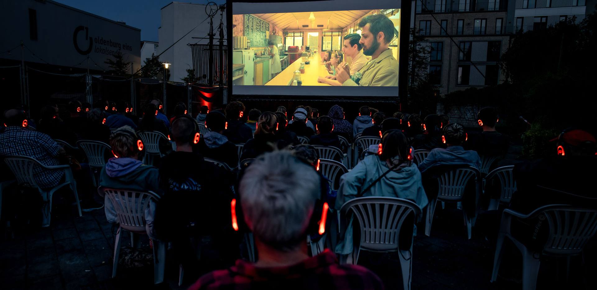 Weite Perspektive mit Blick von hinten auf den Kulturplatz und die Leinwand mittig, dahinter viele Personen aiuf Stüheln mit Kopfhörern. Im Dämmerlicht leuchtet die Leinwand und die Umgebung schimmert dunkel.