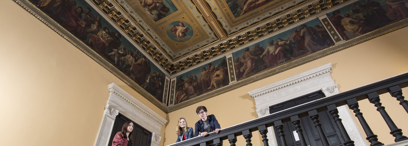 Im Augusteum Oldenburg stehen drei Besucher stehen im historischen Treppenhaus mit Deckengemälde Im Augusteum Oldenburg stehen drei Besucher stehen im historischen Treppenhaus mit Deckengemälde