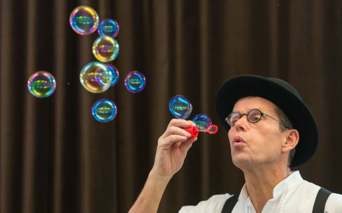 Man mit schwarzem Hut und Brille, der vor einem braunen Vorhang Seifenblasen pustet. Man mit schwarzem Hut und Brille, der vor einem braunen Vorhang Seifenblasen pustet.