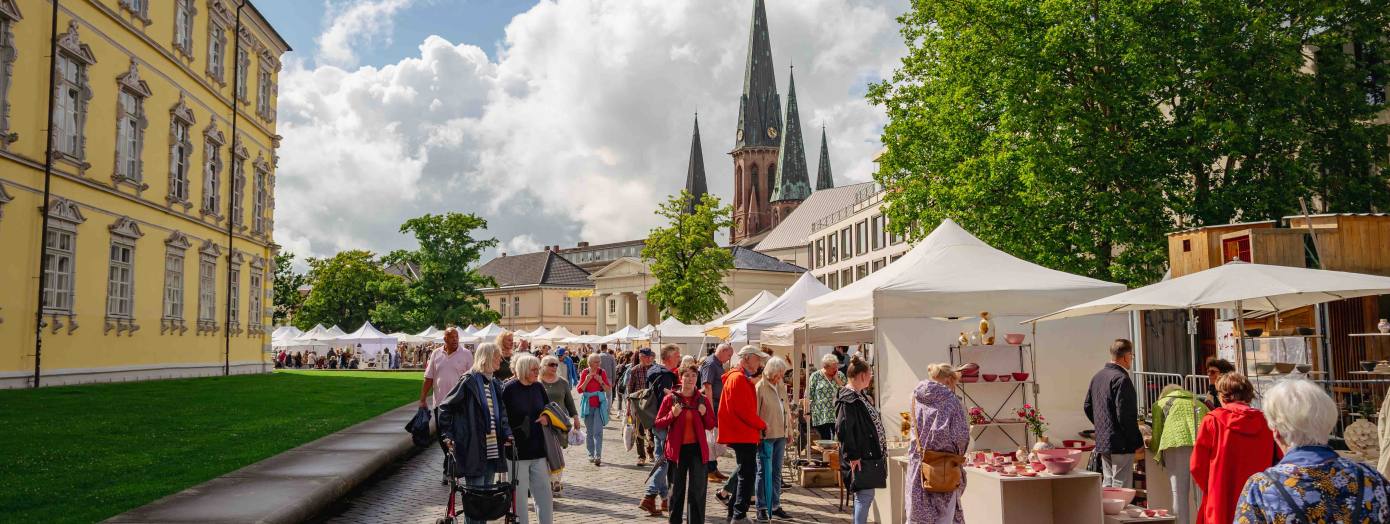 Links sieht man das gelbe Oldenburger Schloss und im fernen Hintergrund die Lambertikirche. Im Vordergrund sieht man verschiedene Menschen an den Keramikmarktständen bummeln. Links sieht man das gelbe Oldenburger Schloss und im fernen Hintergrund die Lambertikirche. Im Vordergrund sieht man verschiedene Menschen an den Keramikmarktständen bummeln.