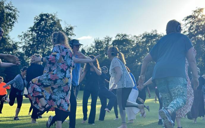 Eine Gruppe von Menschen tanzt auf einer gro&szlig;en Wiese im Freien. Alle Personen haben Kopfh&ouml;rer auf, &uuml;ber die sie die gleiche Musik h&ouml;ren. Die Szene wirkt lebendig und entspannt, w&auml;hrend das warme Gegenlicht der tief stehenden Sonne eine sommerliche Atmosph&auml;re schafft.