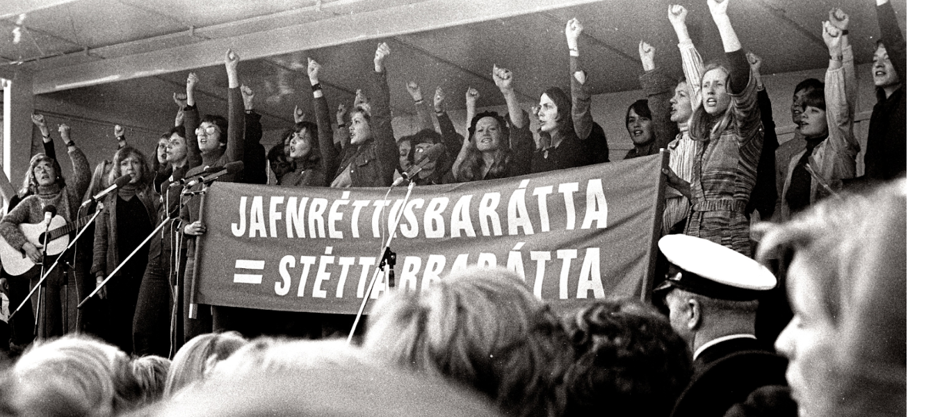 Schwarz weiß Bild mit vielen Frauen die auf einer Bühne stehen, ihre linke Faust heben und einen Banner heben.