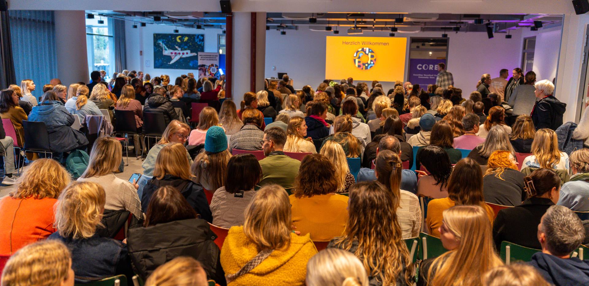 Ein voll besetzter Veranstaltungssaal mit rund 250 Menschen, die auf bunten Stühlen sitzen und nach vorne zur Bühne blicken. Auf einer großen Leinwand steht ‚Herzlich Willkommen' mit dem bunten Logo der Bildungsregion Stadt Oldenburg. Foto: Liz Hussein Ein voll besetzter Veranstaltungssaal mit rund 250 Menschen, die auf bunten Stühlen sitzen und nach vorne zur Bühne blicken. Auf einer großen Leinwand steht ‚Herzlich Willkommen' mit dem bunten Logo der Bildungsregion Stadt Oldenburg. Foto: Liz Hussein