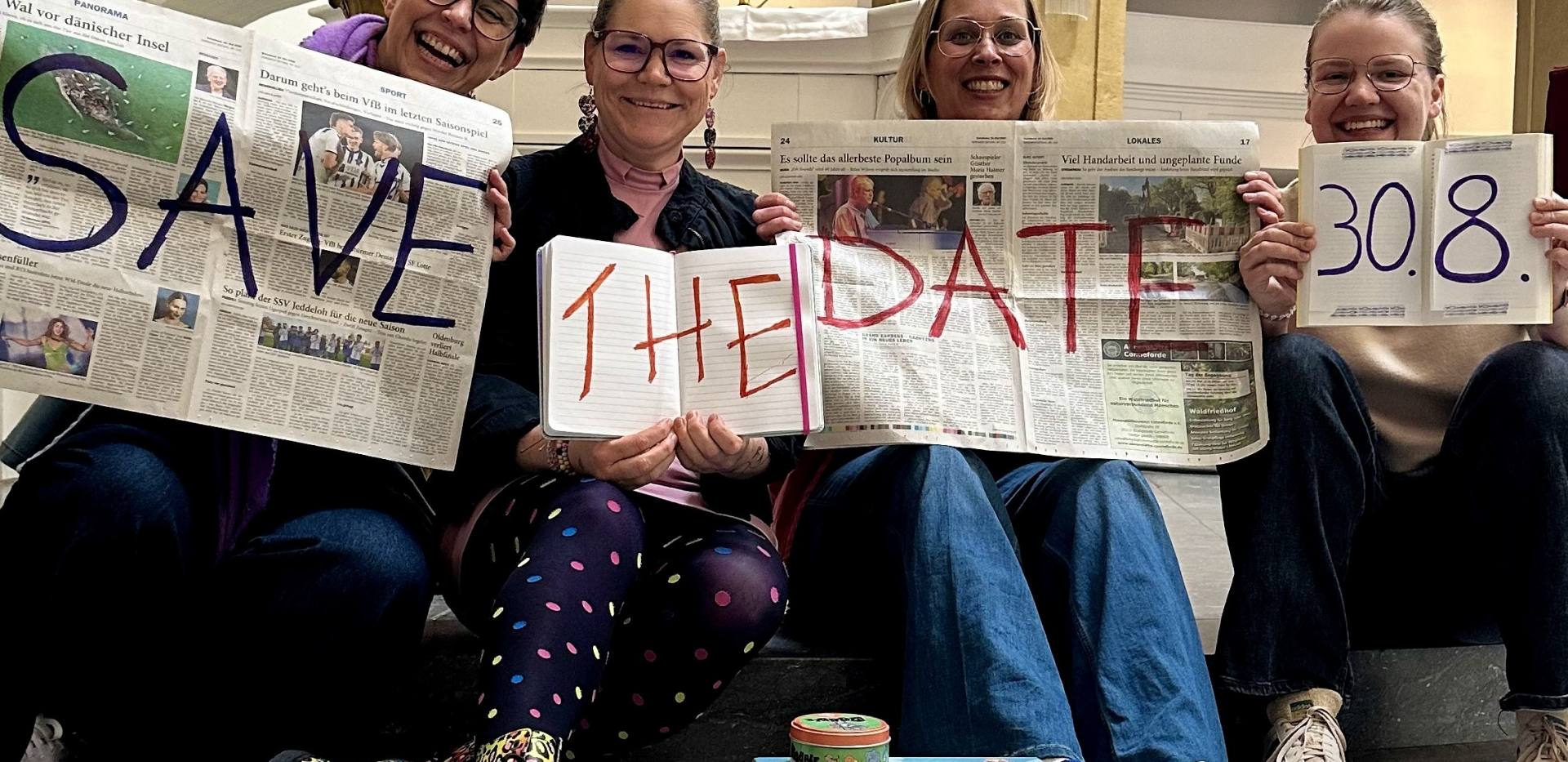 Jenny, Meike, Nele und Gesa halten Zeitung, Heft, Buch hoch mit dem Text "Safe the date, 30.8." Vor ihnen sind Spiele, Bücher, Stifte Jenny, Meike, Nele und Gesa halten Zeitung, Heft, Buch hoch mit dem Text "Safe the date, 30.8." Vor ihnen sind Spiele, Bücher, Stifte