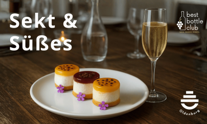 Drei s&uuml;&szlig;e Petits Fours mit essbarer Bl&uuml;te auf einem wei&szlig;en Teller, rechts daneben ein Glas Sekt.