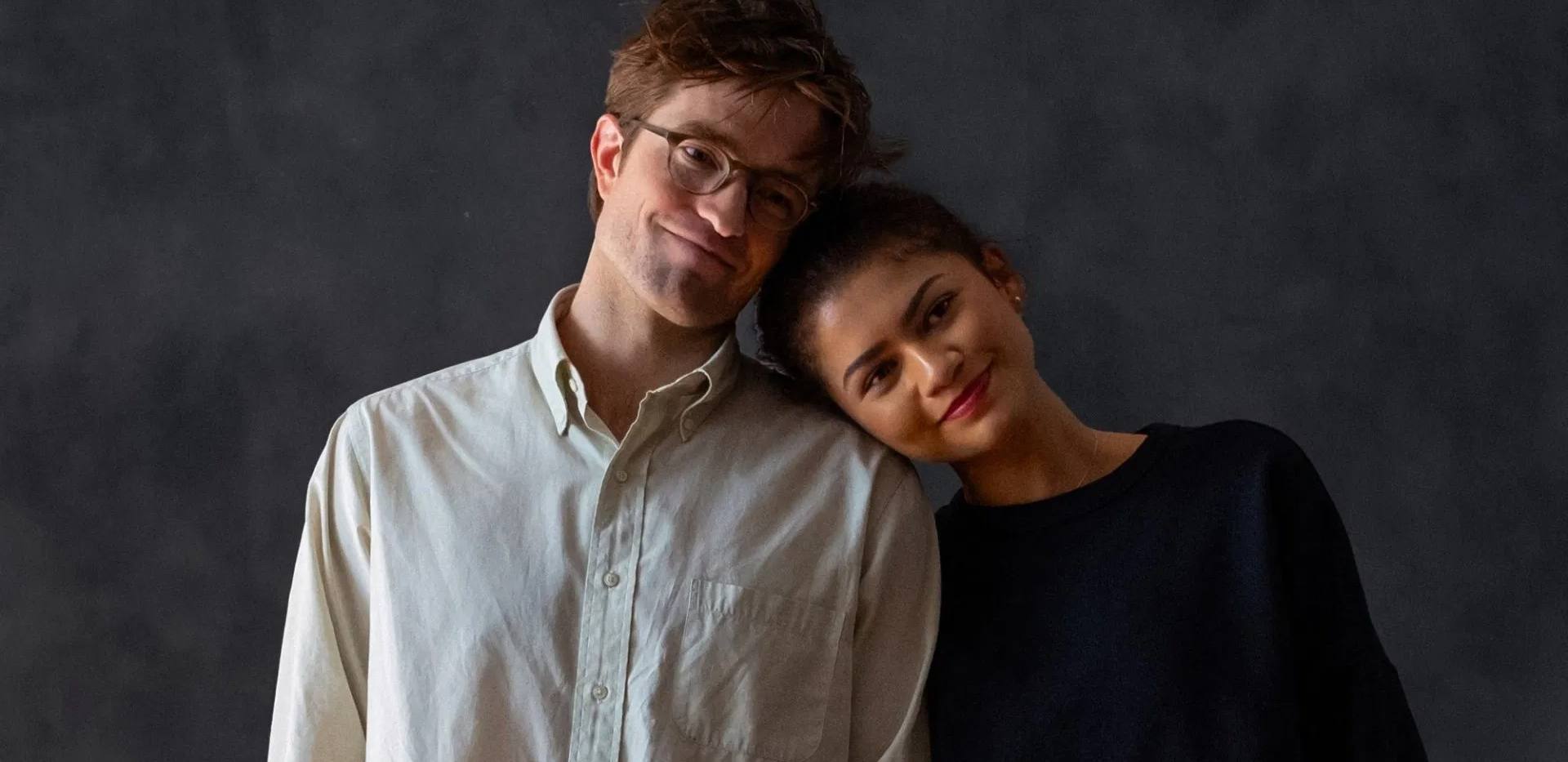Auf dem Bild sind 2 Personen, die vor einem dunklen Hintergrund stehen. Die als Frau gelesene Person rechts lehnt sich an die linke Person, die als Mann gelesen wird. Beide Personen haben einen glücklichen / zufriedenen Gesichtsausdruck. Auf dem Bild sind 2 Personen, die vor einem dunklen Hintergrund stehen. Die als Frau gelesene Person rechts lehnt sich an die linke Person, die als Mann gelesen wird. Beide Personen haben einen glücklichen / zufriedenen Gesichtsausdruck.