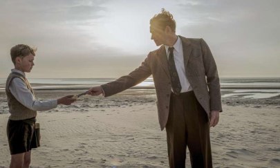 2 Personen stehen bei Sonnenauf- oder Untergang am Strand. Im Hintergrund sieht man das Meer. Eine Person steht rechts und ist erwachsen und die andere Person steht links und ist ein Kind. Die Person rechts &uuml;berreicht der Person links etwas.