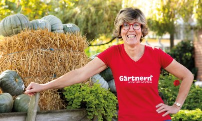 Zu sehen ist Katrin Iskam. Sie tr&auml;gt ein rotes T-shirt worauf G&auml;rtnern ohne viel Geschiss steht. Sie leht ihren rechten Arm auf einem Heuwagen ab.
Auf diese Wagen liegt Stroh und K&uuml;rbisse.
Im Hintergrund sieht man einen Garten