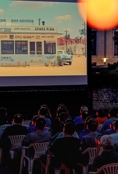 Nähere Perspektive mit seitlichem Blick auf den Kulturplatz und die Leinwand, die fast die Hälfte des Bildes ausfüllt. Davor viele Personen auf Stüheln mit Kopfhörern. Eine orangefarbene Reflektion einer Lichterkette ist im oberen Bildrand zu sehen.