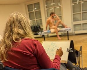 Zeichnende Teilnehmerin des Aktkurses mit Aktmodel im Hintergrund.