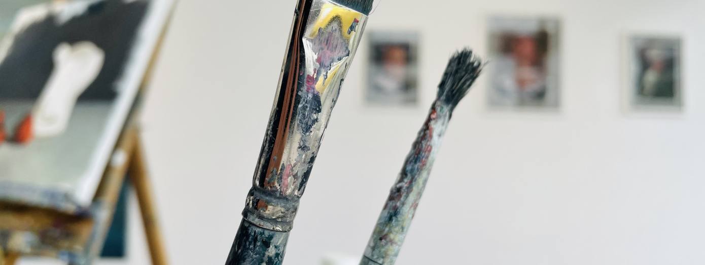 Auf dem Foto befinden sich einige Pinsel, im Hintergrund sieht man Gem&auml;lde.