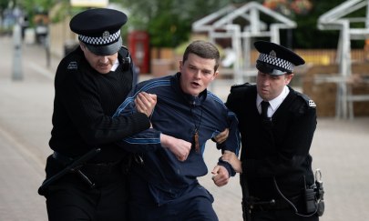 3 Personen stehen auf einer Straße. 2 Personen sind wie Polizisten gekleidet und stehen jeweils links und rechts neben der 3. Person. Die 2 wie Polizisten gekleidete Personen greifen jeweils angestrengt einen Arm der 3. Person und führen sie wohin. 3 Personen stehen auf einer Straße. 2 Personen sind wie Polizisten gekleidet und stehen jeweils links und rechts neben der 3. Person. Die 2 wie Polizisten gekleidete Personen greifen jeweils angestrengt einen Arm der 3. Person und führen sie wohin.