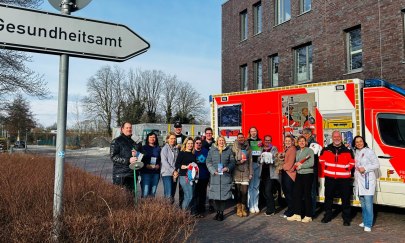 Mitarbeiter des Gesundheitsamtes stehen vor einem Schild "Gesundheitsamt" und einem Rettungswagen.