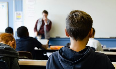 Mehrere Sch&uuml;ler sitzen an Schultischen in Reihen, im Hintergrund steht eine erwachsene Person.