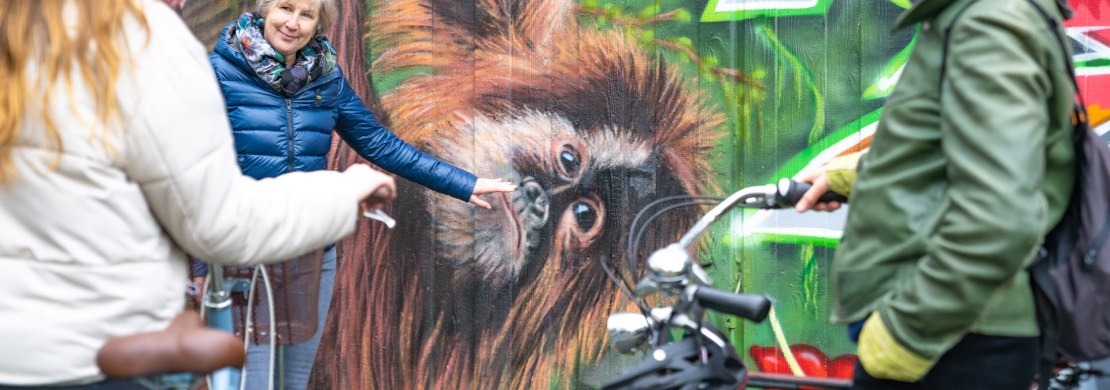 Eine Gästeführerin zeigt zwei Touristen Street Art mit einem Affen in Oldenburg bei einer geführten Radtour