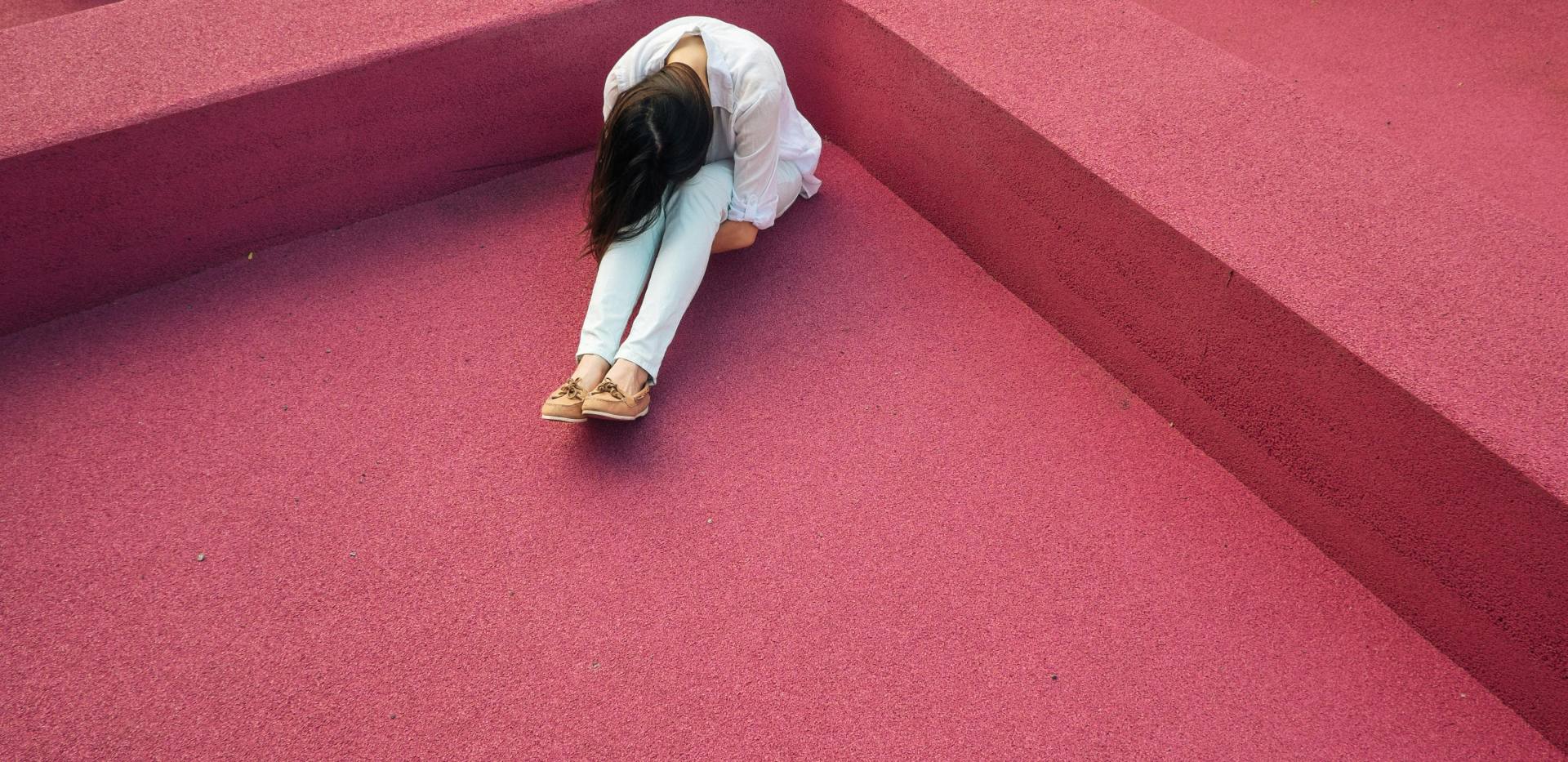 Eine Person mit langen Haaren sitzt auf dem Boden in der Ecke eines Raumes und beugt ihren Oberkörper nach vorn, das Gesicht liegt auf den Knien. Eine Person mit langen Haaren sitzt auf dem Boden in der Ecke eines Raumes und beugt ihren Oberkörper nach vorn, das Gesicht liegt auf den Knien.