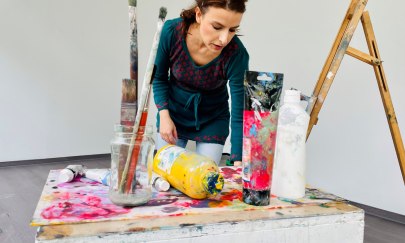 Man sieht die Malerin Milena Tsochkova die vor einer großen weißen Wand steht. Vor ihr stehen verschiedene Farben und Pinsel auf einem Tisch. Auf der rechten Seiten kann man noch eine Leiter sehen.