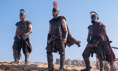 Auf dem Bild sind 3 männlich gelesene Personen zu erkennen, die altertümliche Rüstungen tragen. Die Personen stehen draußen auf rauen Boden und schauen herab. Auf dem Bild sind 3 männlich gelesene Personen zu erkennen, die altertümliche Rüstungen tragen. Die Personen stehen draußen auf rauen Boden und schauen herab.