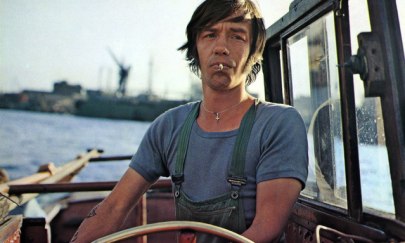 Eine m&auml;nnlich gelesene Person mit Zigarette im Mund man&ouml;vriert ein Boot und steht vor dem Lenkrad. Die Person befindet sich mit dem Boot auf dem Wasser. Im Hintergrund kann man verschwommen einen Hafen erkennen.