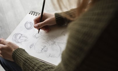 Man sieht eine Person in einem dunklem Pullover die auf einem Block verschiedene Dinge mit einem Bleistift zeichnet. Man sieht eine Person in einem dunklem Pullover die auf einem Block verschiedene Dinge mit einem Bleistift zeichnet.