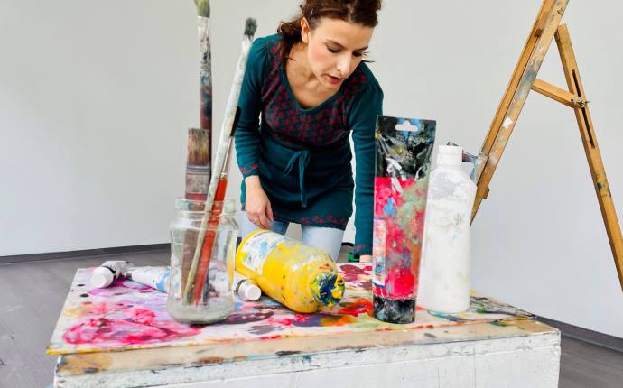 Man sieht die Malerin Milena Tsochkova die vor einer großen weißen Wand steht. Vor ihr stehen verschiedene Farben und Pinsel auf einem Tisch. Auf der rechten Seiten kann man noch eine Leiter sehen.